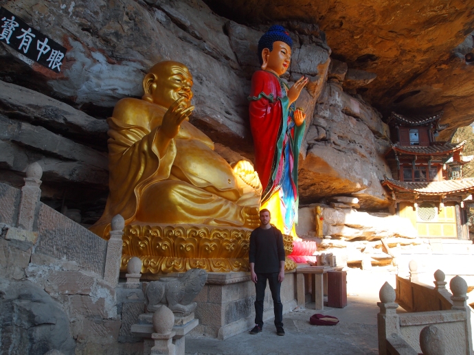 Alex stands beside Guanyin and Maitreya on the cliff ledges at Baoxiang Temple